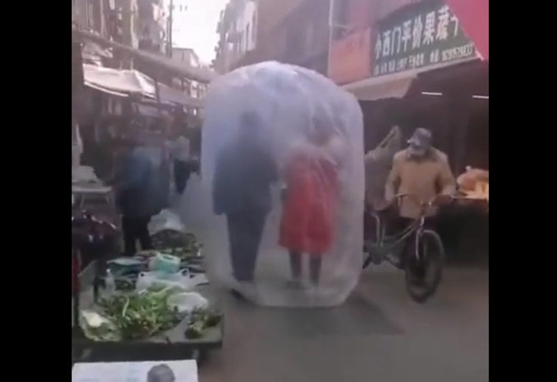 رجل وامرأة يعتمران غلافاً بلاستيكياً شفافاً وكبيراً يحيط بهما خلال تجوالهما في أحد الأسواق الصينية